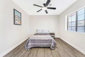 Um quarto com cama, ventilador de teto, janela com persianas e um quadro na parede.