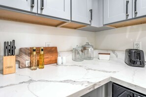 Uma cozinha com armários brancos, bancada de mármore, porta-facas, dois frascos de perfume, saleiro e pimenteiro, potes de vidro, uma torradeira e uma tábua de corte de madeira.