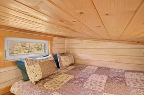 A cabin interior with a wooden ceiling, a window with a view of trees, a bed with patterned bedding, and a small table. - Cozy Teeny Tiny Home with New River view. Pet friendly. (Todd)