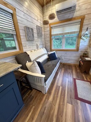 A cozy cabin interior with a wooden floor, a sofa with cushions, a small table, and a window with blinds. - Cozy Teeny Tiny Home with New River view. Pet friendly. (Todd)