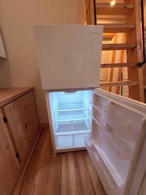 Een witte koelkast met houten binnenkant, een houten kastje links en een houten trap op de achtergrond.