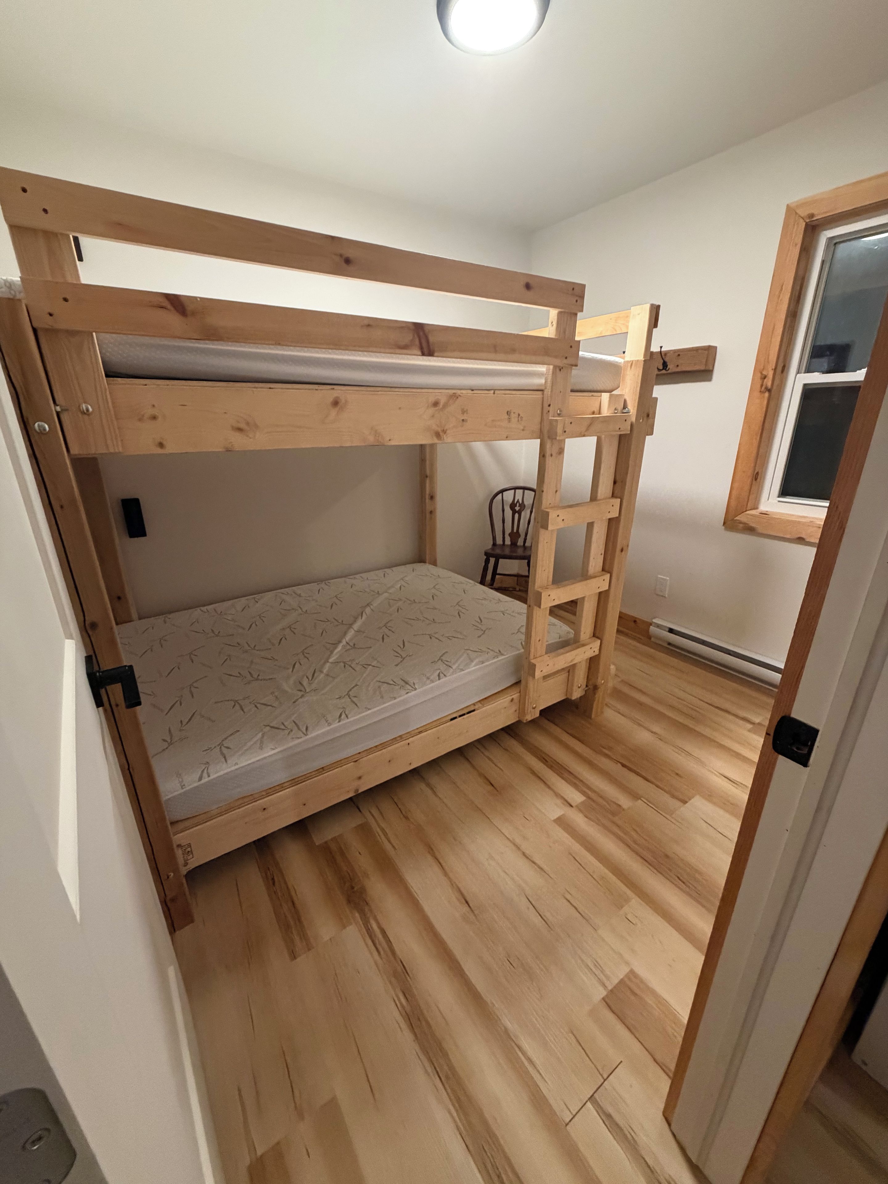 A bunk bed with a wooden frame, a mattress with a leaf pattern, and a chair in a room with wooden flooring and a window.
