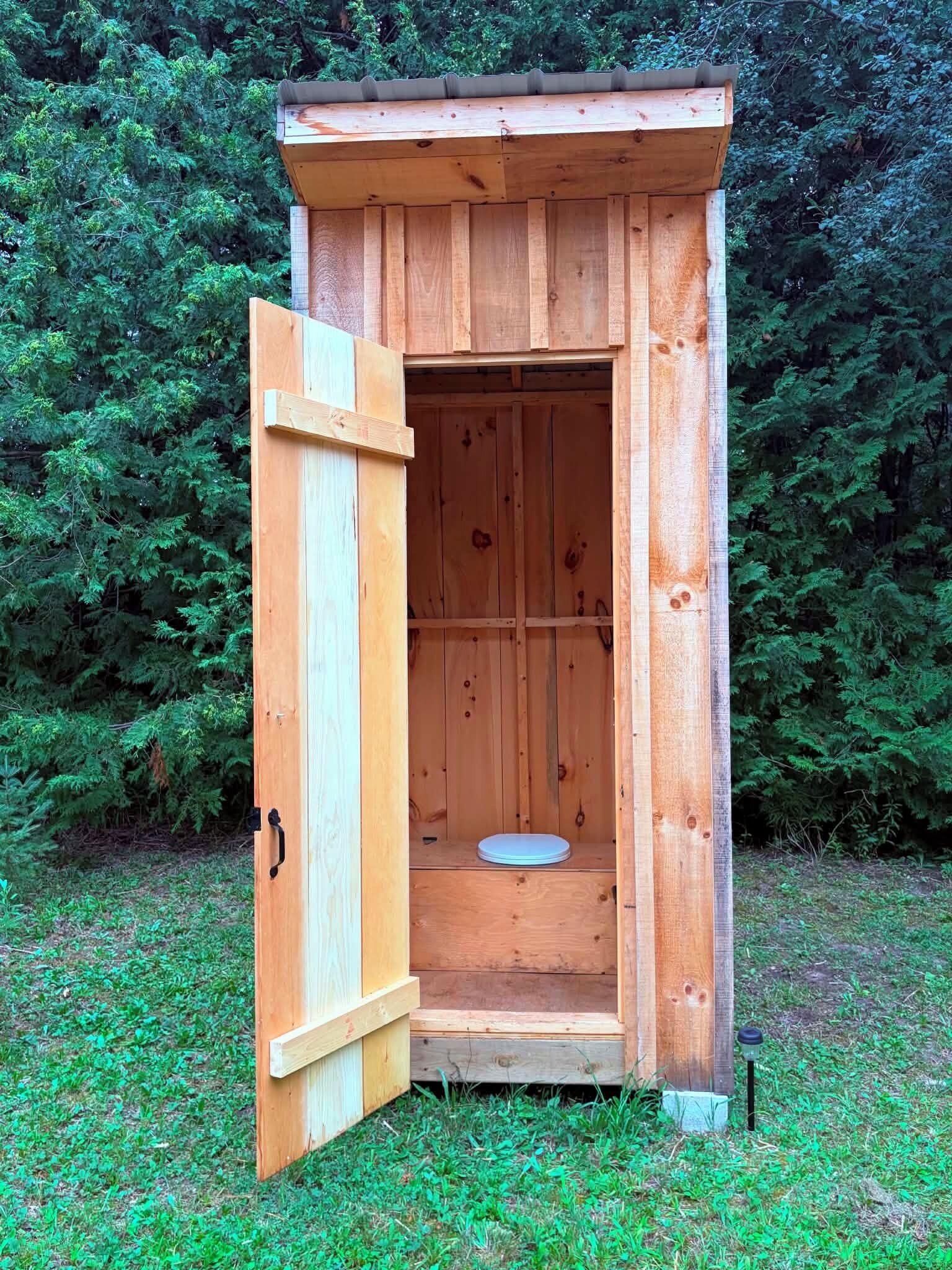 Eine hölzerne Gartentoilette, deren Tür offen steht und einen Blick auf die Toilette im Inneren freigibt.