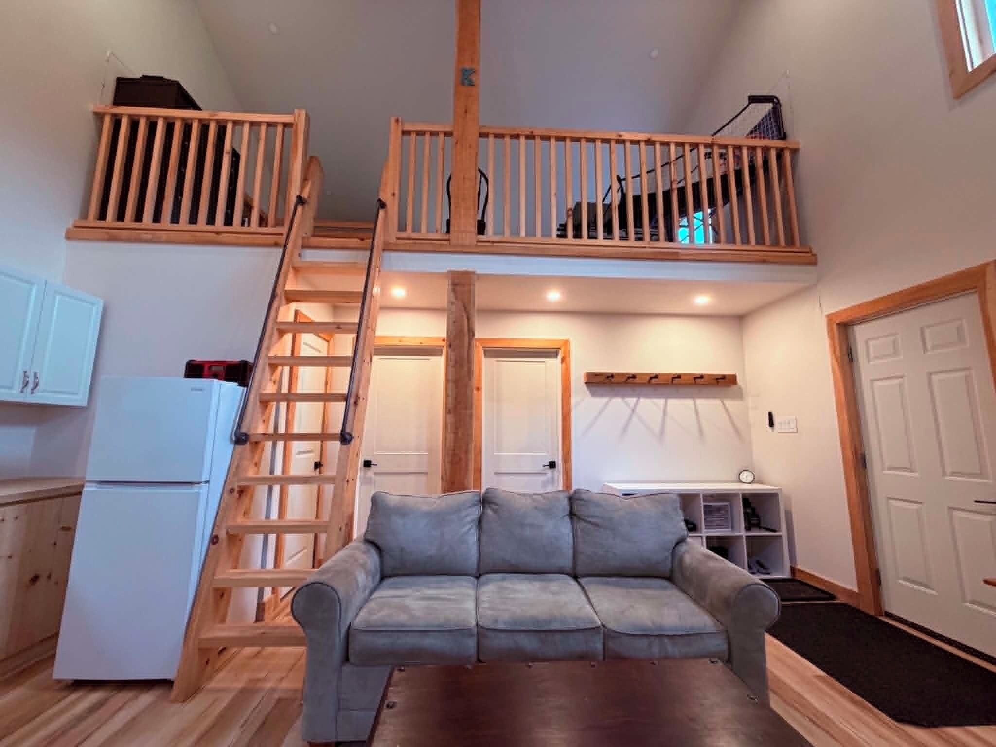 A loft-style living space with a wooden staircase, a sofa, a kitchenette, and a small dining area.