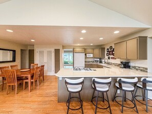 A modern kitchen with a central island, bar stools, and a dining area with wooden chairs. - Spacious McMinnville Retreat | 5 Bedrooms | Pet Friendly (Mcminnville)