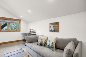 Ein Wohnzimmer mit grauem Sofa, zwei Zierkissen, einem Bild in Holzrahmen und einem Fenster mit Jalousien.