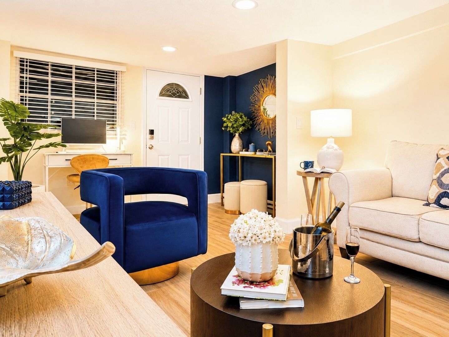 Een moderne woonkamer met een blauwe fauteuil, een beige bank, een salontafel met een boek en een decoratieve vaas, en een televisie op de achtergrond.