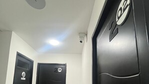 Un pasillo con dos puertas negras, una con un cartel que indica '101' y otra con '102', y una cámara de seguridad en el techo.