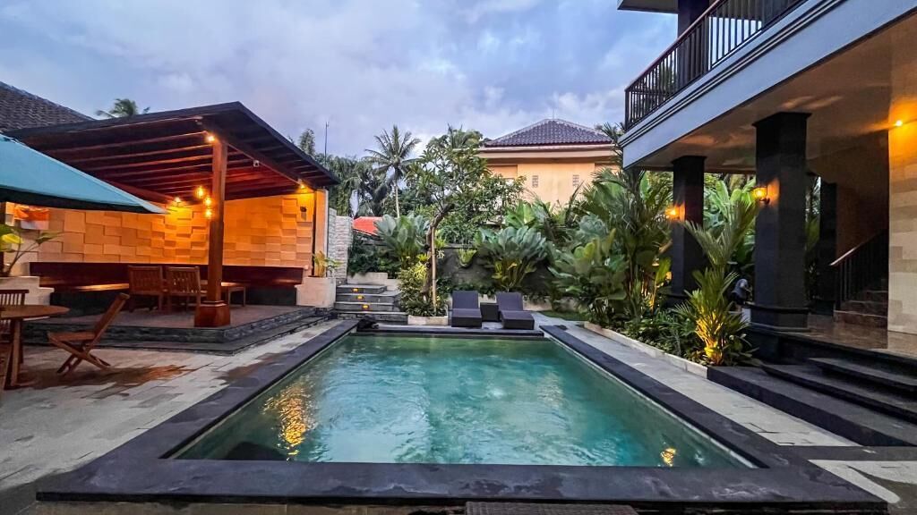 Uma piscina com borda de pedra, rodeada por uma área coberta com assentos e uma escadaria que leva a um edifício.