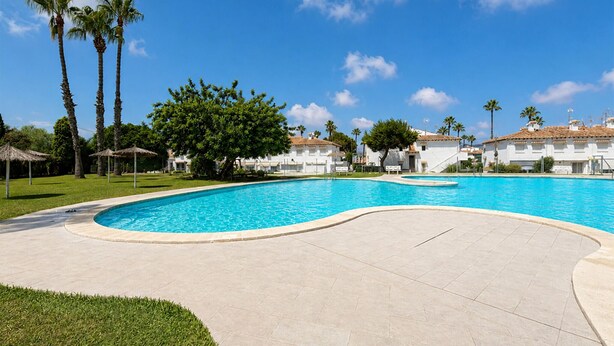 Una piscina de borde curvo, rodeada de una zona pavimentada, palmeras y edificios blancos con techos de tejas rojas.