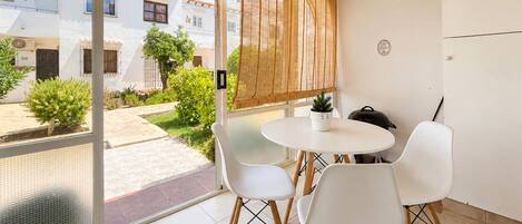 Un balcón con una mesa y sillas blancas, una planta en maceta y vistas a un patio con edificios.