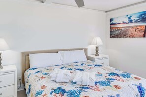 Ein ordentlich bezogenes Bett mit einer blumigen Tagesdecke, zwei weiße Kissen und gefaltete Handtücher auf einem weißen Nachttisch.