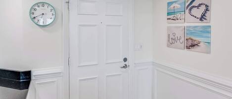 Un pasillo con una puerta blanca, un reloj de pared y tres cuadros enmarcados.
