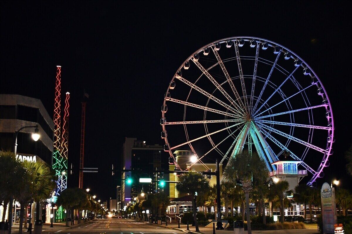 A large Ferris wheel with a colorful light display, a tall illuminated tower, and a street lined with palm trees and buildings.