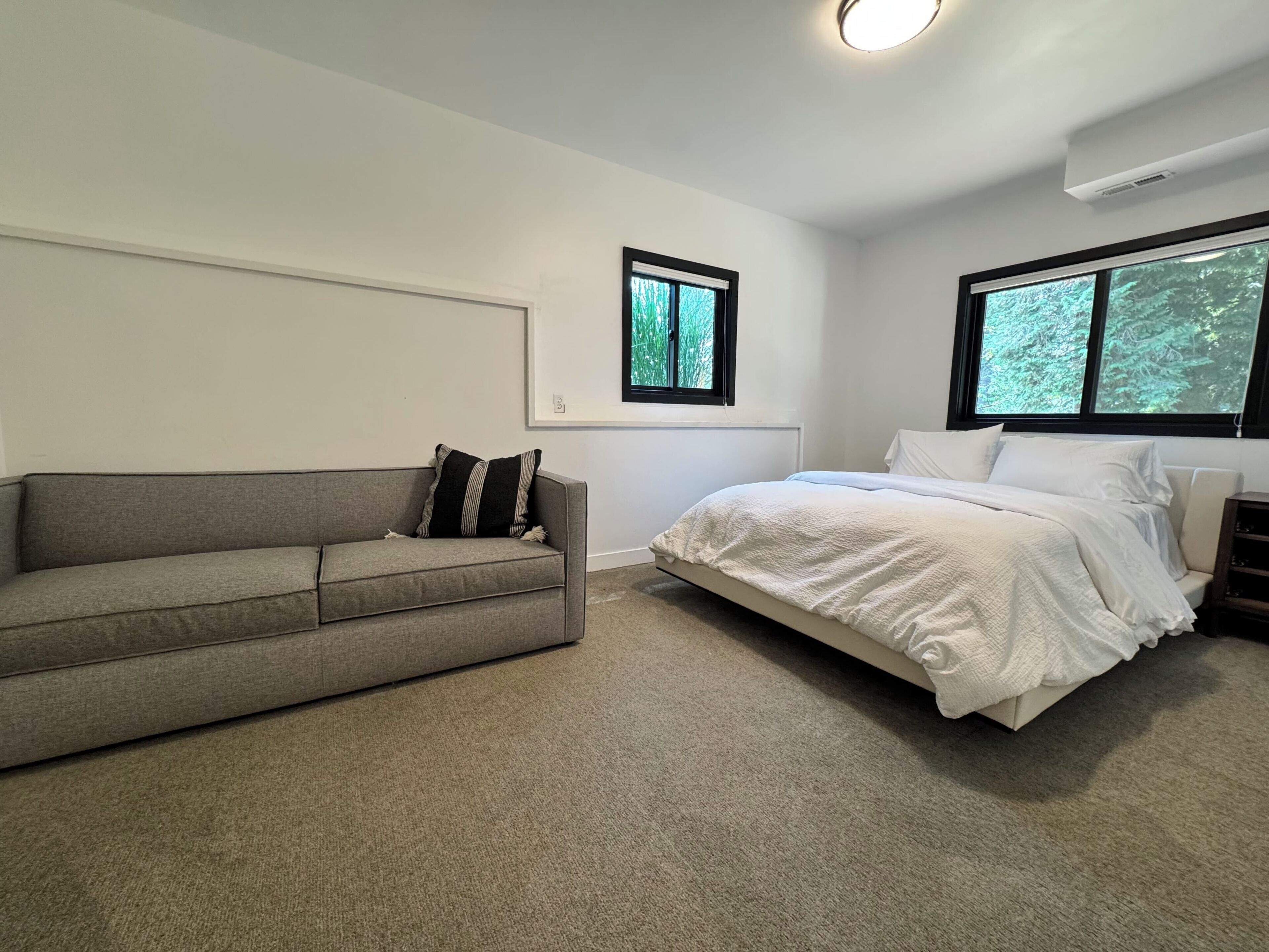 A bedroom with a grey sofa, a white bed, and two windows with views of greenery.