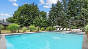 Ein rechteckiger Swimmingpool mit einer gefliesten Kante, umgeben von einem Metallzaun, und ein Liegebereich mit weißen Stühlen und einem Sonnenschirm.