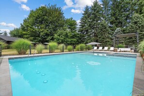 A rectangular swimming pool with a tiled edge, surrounded by a metal fence, and a lounge area with white chairs and a shaded umbrella. - Modern Luxe Estate~Pool·HotTub·2AcresPrivateResort (Issaquah)