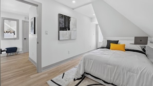 A modern bedroom with a sloped ceiling, a bed with white bedding and a yellow pillow, a desk with a laptop, and artwork on the walls.