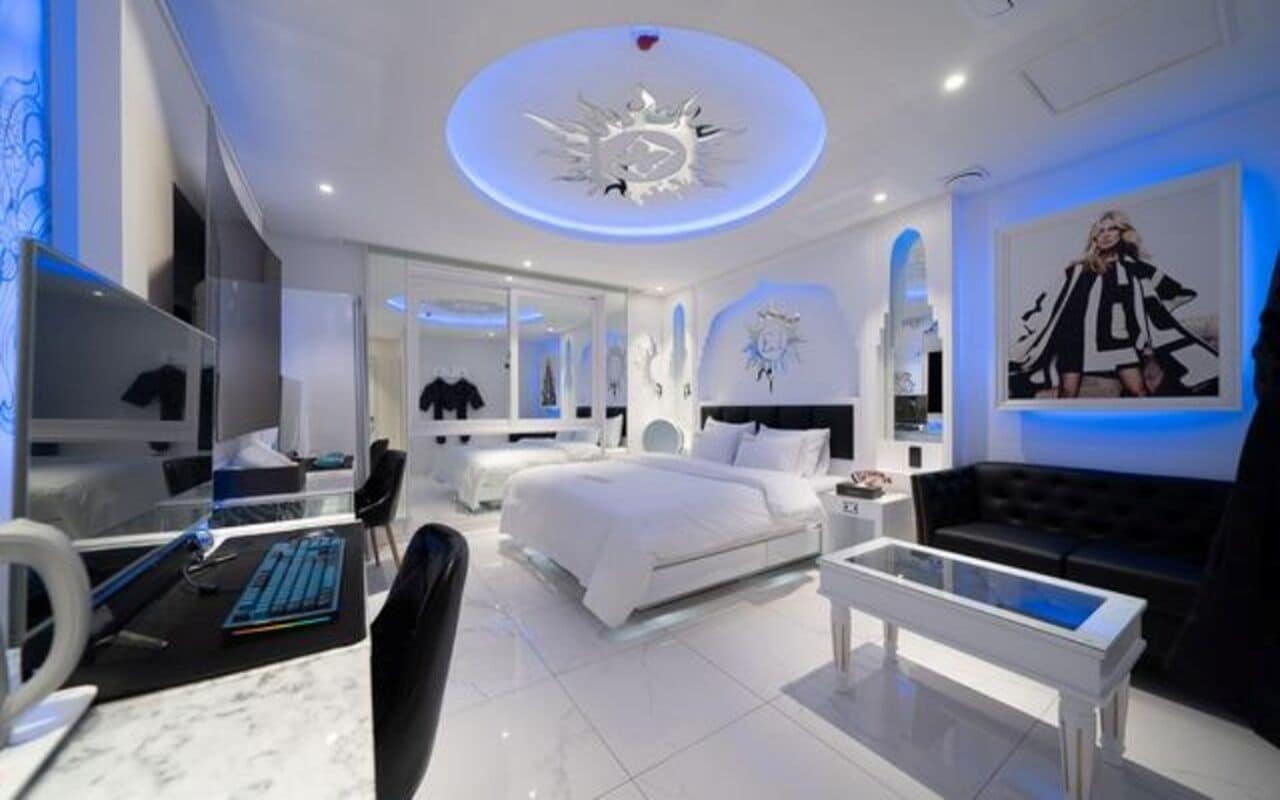A modern hotel room with a bed, a desk with a computer, a sofa, and a large mirror.