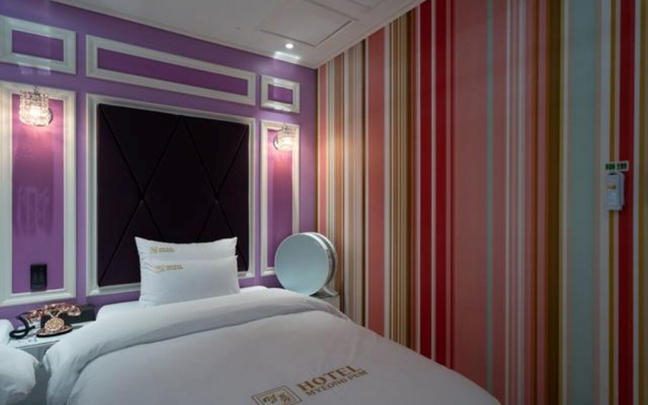A hotel room with a large bed, a headboard with a diamond pattern, a bedside table with a lamp, and a striped wall.