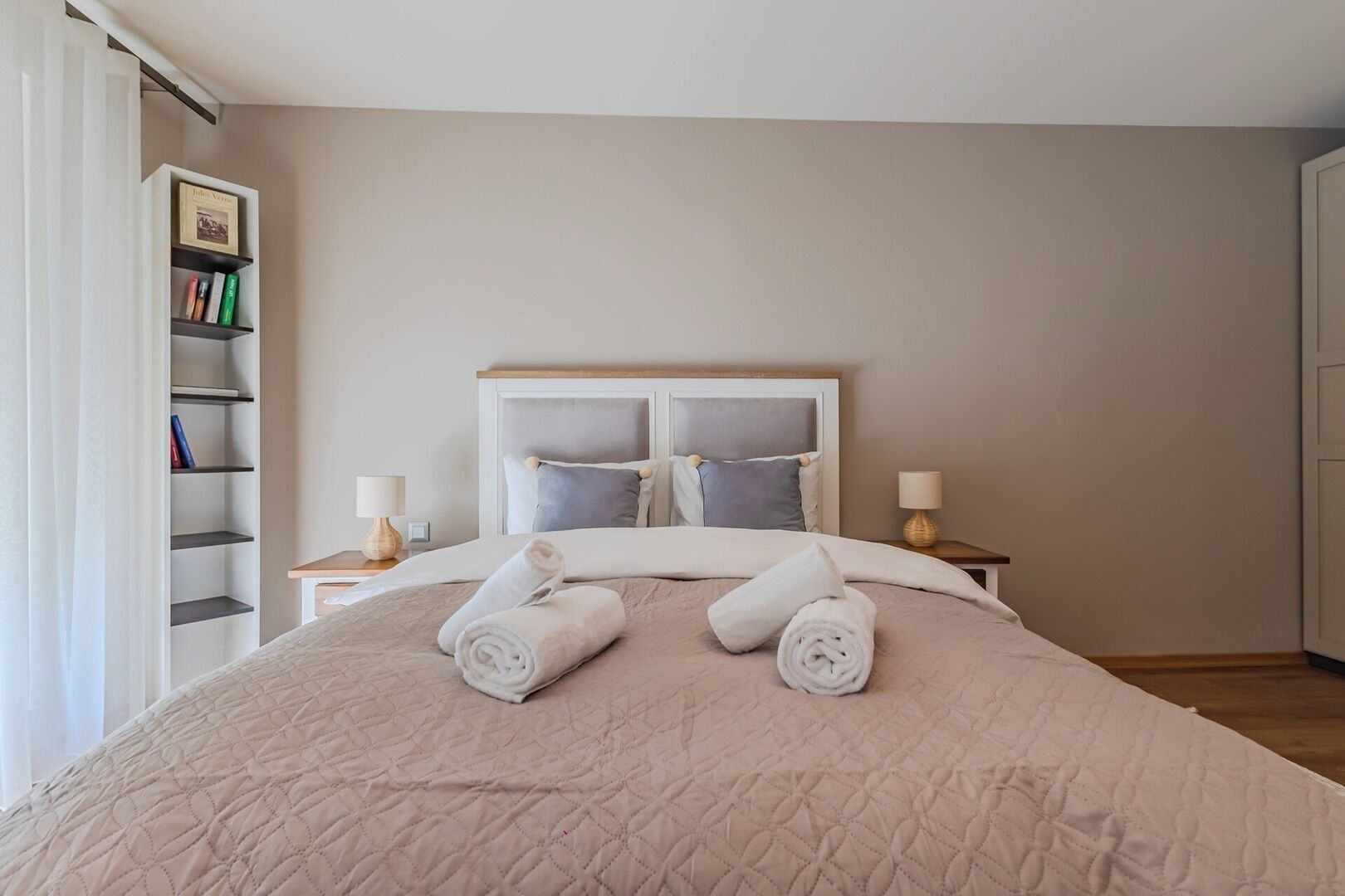 A neatly made bed with rolled towels, a bookshelf with books, and two bedside lamps.