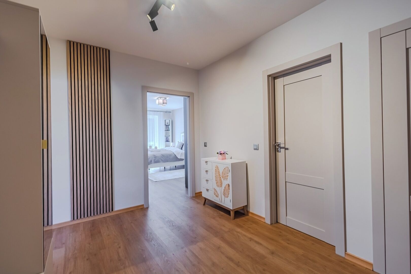 Un corridoio con pavimento in legno, una porta bianca e un comodino con un oggetto decorativo.