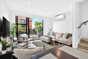 Ein modernes Wohnzimmer mit Sofa, Sessel und Couchtisch, einem Flachbildfernseher und Blick auf ein Backsteinhaus durch große Fenster.