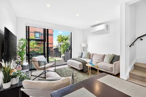 A modern living room with a sofa, armchair, and coffee table, a flat-screen TV, and a staircase leading to an upper floor. - Serene 3BR Home Close to City & Parks Zodiak Stays (Auckland)