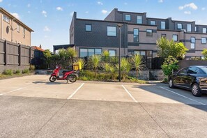 Ein Parkplatz mit einem roten Roller, einem schwarzen Auto und einem Gebäude mit vielen Fenstern und einer Ziegelwand.