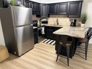 A modern kitchen with dark cabinetry, a stainless steel refrigerator, a microwave, a coffee maker, and a granite countertop. - Perfect Walkable Midtown Location, Ocean View, Spacious 2 BR 2 Bath Condo (Ocean City)