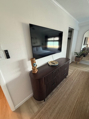 A modern living room with a flat-screen TV mounted on a dark wooden console, a decorative wooden sculpture, and a woven rug on the floor. - Perfect Walkable Midtown Location, Ocean View, Spacious 2 BR 2 Bath Condo (Ocean City)