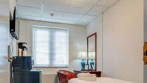 Une chambre d’hôtel comprenant un lit, un bureau avec un ordinateur, une chaise, une fenêtre avec des stores, une lampe et une machine à café.