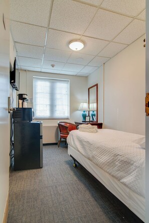 A hotel room with a bed, a desk with a computer, a chair, a window with blinds, a lamp, and a coffee machine. - Historic Converted Dorm Room off Vanderbilt! (Nashville)