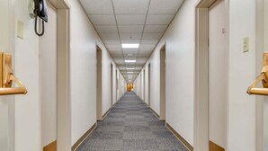 Un long couloir étroit, typique des hôtels, avec des portes des deux côtés, un revêtement de sol en moquette et un plafond éclairé par des néons.