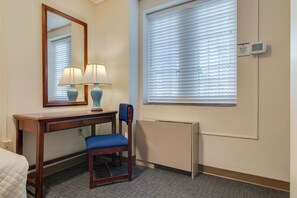 Ein Hotelzimmer mit Schreibtisch aus Holz, zwei Lampen, einem Sessel, einem großen Spiegel, einem Fenster mit Jalousien und einem Thermostat.