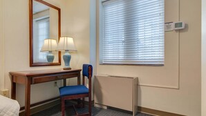 Hotellihuone, jossa on puinen työpöytä, kaksi lamppua, tuoli, suuri peili, ikkuna, jossa on sälekaihtimet, ja termostaatti seinässä.
