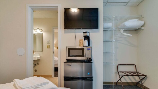Quarto de hotel com cama, televisão, máquina de café, micro-ondas, prateleira com toalhas e uma cadeira.