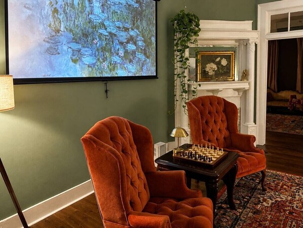 A room with two armchairs, a chessboard, a painting, and a fireplace.