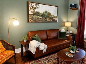 A living room with a brown leather sofa, a patterned armchair, a round coffee table, a painting on the wall, and a lamp on a side table.