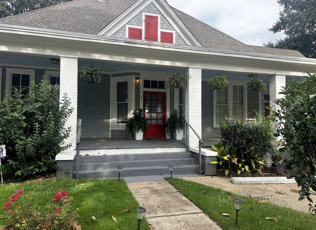 Una casa a due piani con un portico, una porta rossa e un piccolo giardino antistante con un vialetto che conduce all'ingresso.