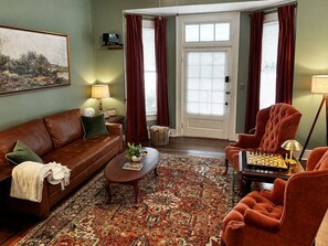 A living room with a brown leather sofa, a coffee table, a patterned rug, a chess table, and a door with red curtains.