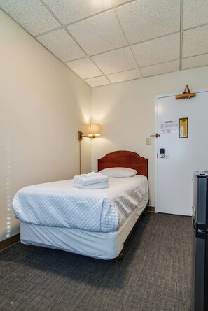 Ein Hotelzimmer mit einem Einzelbett, einem Nachttisch, einer Lampe, einer Tür mit Schloss und einem kleinen Kühlschrank.