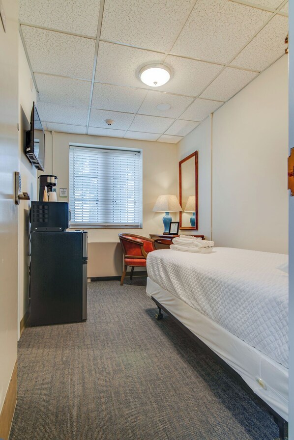 Ein Hotelzimmer mit Bett, Schreibtisch, Stuhl, Fernseher, einem Fenster mit Jalousien und einer Deckenleuchte.