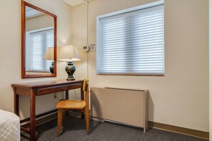Ein Hotelzimmer mit Schreibtisch aus Holz, einem Stuhl, einer Lampe, einem Spiegel, einem Fenster mit Jalousien und einer Heizung.