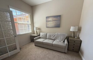 Ein Zimmer mit einem beige gefärbten Sofa, einem kleinen Beistelltisch mit Lampe, einem Bild an der Wand und einer Tür mit satinierten Glasscheiben.