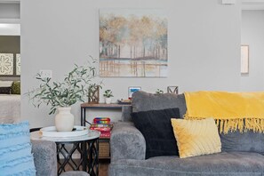 Ein Wohnzimmer mit grauem Sofa, gelbener Decke, weißer Vase mit Grünpflanzen und einem Gemälde einer Winterlandschaft.