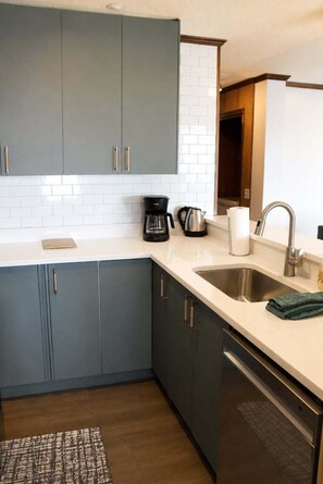 Eine moderne Küche mit grauen Schränken, einer weißen Arbeitsplatte, einer Kaffeemaschine, einem Wasserkocher und einem Edelstahl-Spülbecken.