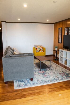 Ein Wohnzimmer mit grauem Sofa, einem gelben Sessel, einem Couchtisch und einer TV-Konsole mit Regalen.