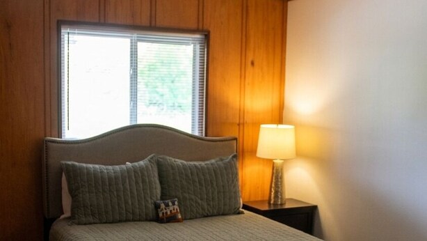 Un dormitorio con cama, mesita de noche, lámpara y ventana.