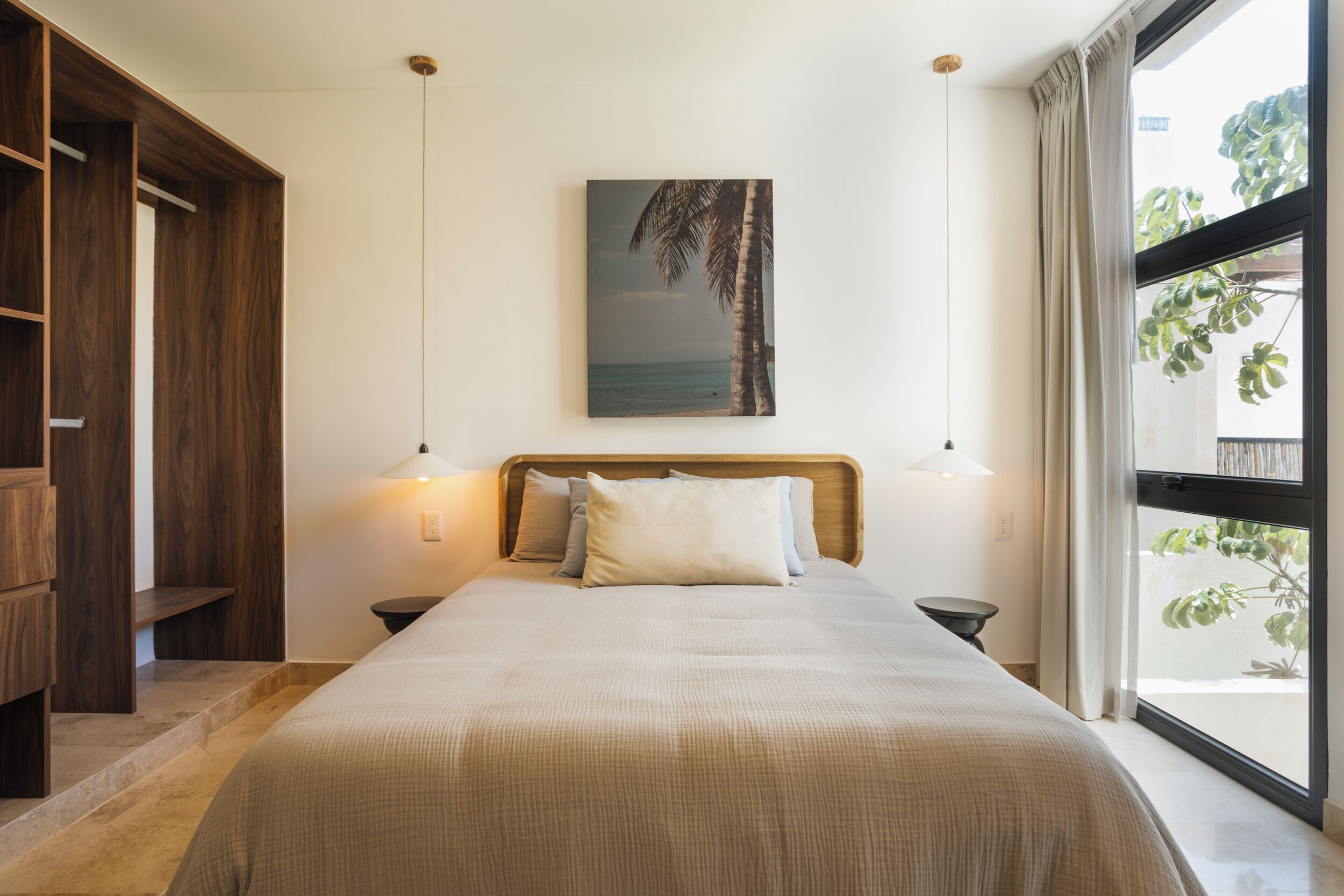 A bedroom with a bed, bedside lamps, a wooden wardrobe, a painting, and a window with curtains.